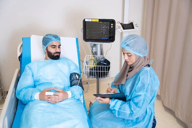 جهاز مزود بشاشة سوداء تعرض بيانات المريض الحيوية مثل النبض وضغط الدم,  طبيبة عربية خليجية اماراتية محجبة تقوم بقراءة الفحوصات, دمج التكنولوجيا الحديثة في المجال الطبي