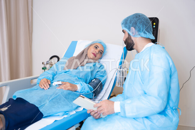 Providing consultations and treatment services, a modern room for examining and caring for patients in the hospital, an Arab Gulf Emirati doctor wearing a medical apron and reading the results of examinations, a veiled patient lying on a hospital bed listening to the doctor
