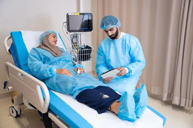 غرفه حديثة لفحص ورعاية المرضى في المستشفى، طبيب عربي خليجي اماراتي يرتدي المريول الطبي و يقوم بقراءة نتيجة الفحوصات, مريضة محجبة مستلقية على سرير المشفى تنصت للطبيب