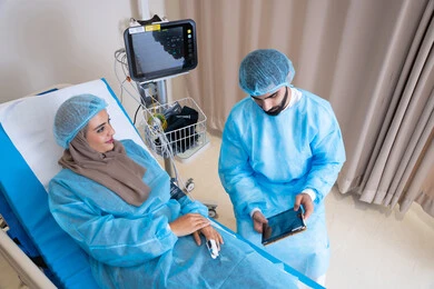 قياس العلامات الحيوية في المستشفى, طبيب عربي خليجي اماراتي يرتدي المريول الطبي و يقوم بقراءة نتيجة الفحوصات عبر التابلت, مريضة محجبة مستلقية على سرير المشفى تنصت للطبيب