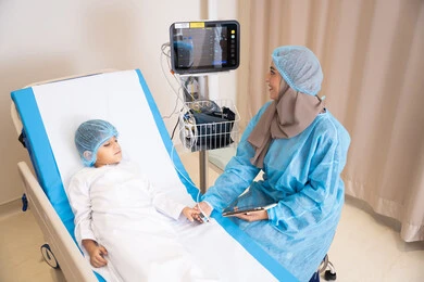 Looks full of optimism, hope and smiles, providing medical consultations in hospitals, health care and treatment, a veiled Arab Gulf Emirati female doctor wearing a medical apron and holding the hand of a sick boy
