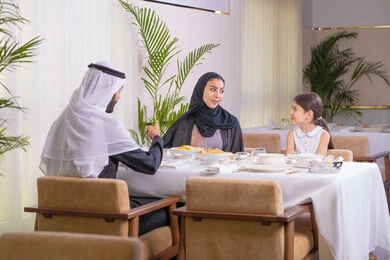 التسامر والدردشة وقضاء وقت ممتع مع العائلة، عائلة عربية خليجية إماراتية يقومون بتناول الطعام، قضاء وقت عائلي ممتع في مطعم فاخر، طاولة مليئة بأطباق الأطعمة الغربية، أجواء أسرية مليئة بالألفة والمحبة، الاستمتاع في عطلة نهاية الأسبوع، خلفية نباتات خضراء