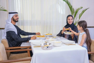 التسلية والترفيه بالعطلات، صورة مقربة لعائلة عربية خليجية إماراتية يقومون بتناول الطعام تقدم الأم الطعام لطفلتها، قضاء وقت عائلي ممتع في مطعم فاخر، طاولة مليئة بأطباق الأطعمة الغربية، أجواء أسرية مليئة بالألفة والمحبة، الاهتمام بالغذاء وصحة الأطفال