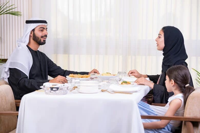 تعزيز التواصل بين الكبار والصغار، صورة من الجانب لعائلة عربية خليجية إماراتية يقومون بالتحدث على طاولة الطعام، أهمية الترابط الأسري والاجتماع العائلي، طاولة مليئة بأطباق الأطعمة الغربية، قضاء وقت ممتع في مطعم راقي