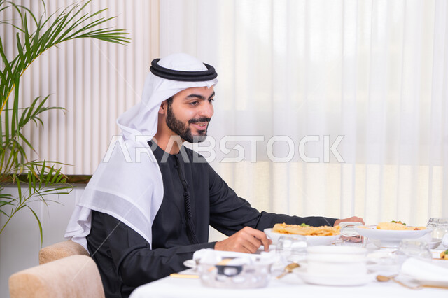 ايماءات السعادة و السرور، رجل عربي خليجي اماراتي يرتدي الكندورة والغترة يقضي وقت ممتع في مطعم راقي، مائدة مليئة بأطباق الأطعمة الشهية، الكرم العربي والأجواء الاجتماعية الودية، التسلية والترفيه في العطلة