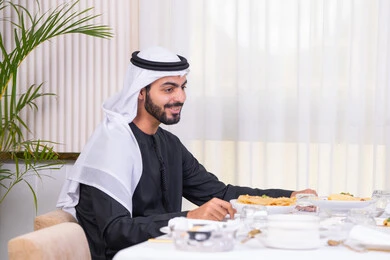 Gestures of happiness and pleasure, an Arab Gulf Emirati man wearing a kandura and ghutra having a good time in a high-end restaurant, a table full of delicious food dishes, Arab generosity and a friendly social atmosphere, entertainment and recreation on holiday