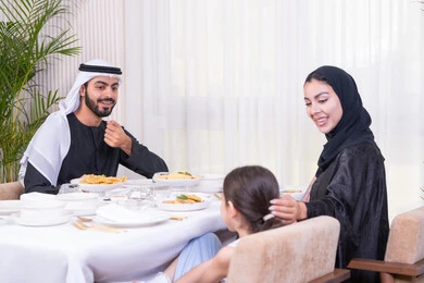 Feelings of love and tenderness in a happy family gathering, an Emirati Gulf Arab family eating with gestures of joy and happiness, spending enjoyable family time in a luxurious restaurant, a table full of Western food dishes, a family atmosphere full of intimacy and love, enjoying the weekend