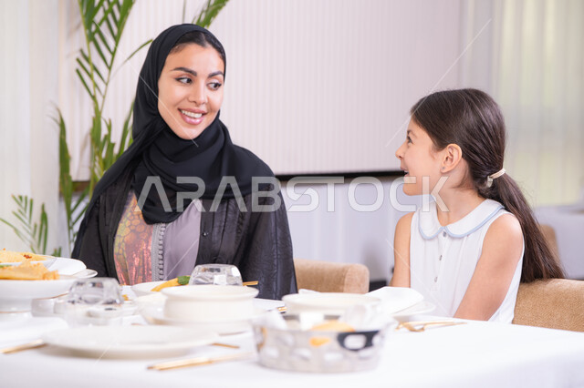 التفاهم والترابط بين الأم والطفلة، فتاه عربية خليجية اماراتية ترتدي الزي الكاجوال رفقة والدتها يقومون بتناول الطعام في مطعم راقي، تقديم الرعاية والحب للأبناء، امرأة عربية خليجية سعودية محجبة ترتدي العباءة السوداء