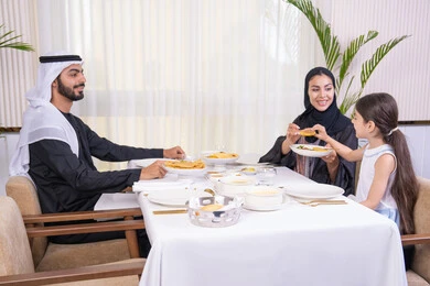 مفهوم العائلة السعيدة، عائلة عربية خليجية إماراتية يقومون بتناول الطعام بإيماءات الحب والسرور،  وضع الأطفال في بيئة عائلية مليئة بالألفة والمشاركة، طاولة مليئة بأطباق الأطعمة الغربية، الاستمتاع في عطلة نهاية الأسبوع، الذكريات واللحظات العائلية الدافئة