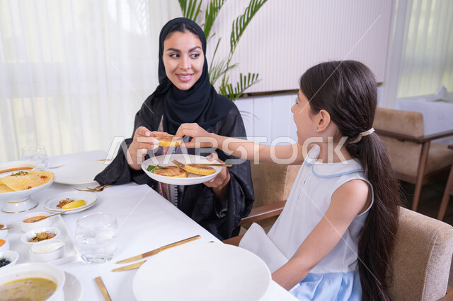التفاهم والترابط بين الأم والطفلة، فتاه عربية خليجية اماراتية رفقة والدتها يقومون بتناول الطعام في مطعم راقي، تقديم الرعاية والمحبة للأبناء، امرأة عربية خليجية سعودية محجبة ترتدي عباءة سوداء تقوم بسكب الطعام لابنتها