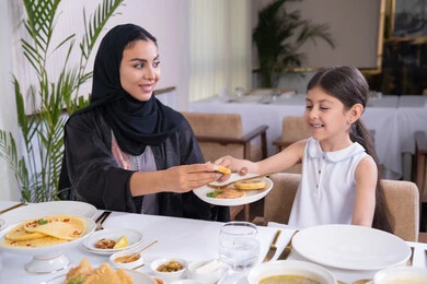 التفاهم والترابط بين الأم والطفلة، فتاه عربية خليجية اماراتية رفقة والدتها يقومون بتناول الطعام في مطعم راقي، تقديم الرعاية والمحبة للأبناء، امرأة عربية خليجية سعودية محجبة ترتدي عباءة سوداء تقوم بسكب الطعام لابنتها