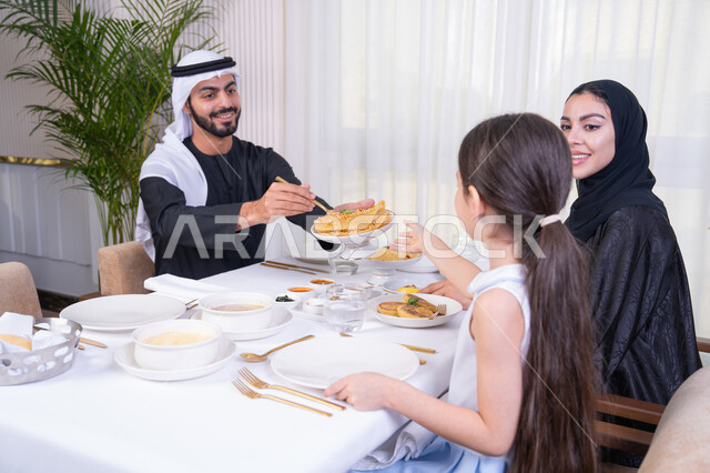 اهتمام الآباء بالأبناء، الاستمتاع في عطلة نهاية الأسبوع، عائلة عربية خليجية إماراتية يقومون بتناول الطعام بإيماءات الحب والسرور،  وضع الأطفال في بيئة عائلية مليئة بالألفة والمشاركة،  الذكريات واللحظات العائلية الدافئة، طاولة مليئة بأطباق الأطعمة الغربية