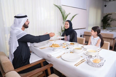 أب عربي خليجي اماراتي يقوم بسكب الطعام لابنته، عائلة عربية خليجية إماراتية يقومون بتناول الطعام بإيماءات الحب والسرور، طاولة مليئة بأطباق الأطعمة الغربية الشهية، الاستمتاع مع الاسرة في عطلة نهاية الأسبوع