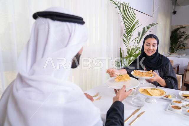 مشاركة الاطعمة بين الزوجين، مائدة مليئة باطباق متنوعه ومختلفه من المأكولات الشهية، الألفة والمودة بين الزوجين، قضاء وقت ممتع خارج المنزل في عطلة نهاية الاسبوع, زوجان عربيان اماراتيان يقومان بتناول الطعام في المطعم, مفهوم الاسره السعيده