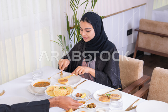 طاولة مليئة باطباق متنوعه ومختلفه من المأكولات الشهية، قضاء وقت ممتعه خارج المنزل في عطلة نهاية الاسبوع, سيدة عربية خليجية اماراتية محجبة ترتدي العباءة تقوم بتناول الطعام في المطعم برفقة زوجها, مفهوم الاسره السعيده