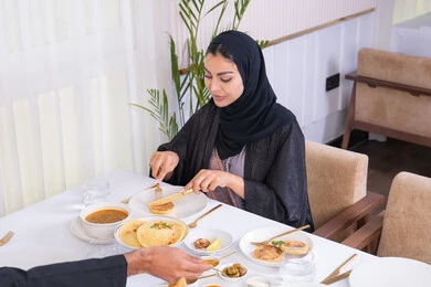 A table full of various delicious dishes, having a good time outside the home on the weekend, a veiled Emirati Gulf Arab woman wearing an abaya eating at a restaurant with her husband, the concept of a happy family