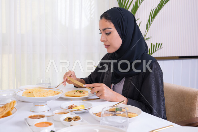 مطاعم راقية في الامارات، طاولة مليئة باطباق متنوعه ومختلفه من المأكولات الشهية، قضاء وقت ممتعه خارج المنزل في عطلة نهاية الاسبوع, صورة من الجانب لامرأة عربية خليجية اماراتية محجبة ترتدي العباءة تقوم بتناول الطعام في المطعم