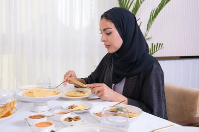 مطاعم راقية في الامارات، طاولة مليئة باطباق متنوعه ومختلفه من المأكولات الشهية، قضاء وقت ممتعه خارج المنزل في عطلة نهاية الاسبوع, صورة من الجانب لامرأة عربية خليجية اماراتية محجبة ترتدي العباءة تقوم بتناول الطعام في المطعم
