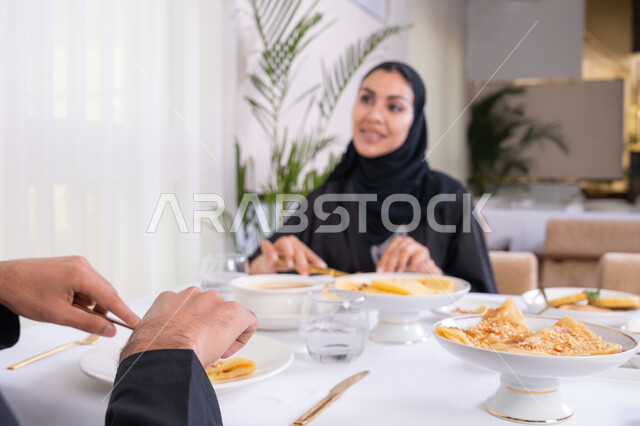 طاولة مليئة باطباق متنوعه ومختلفه من المأكولات الشهية، قضاء وقت ممتعه خارج المنزل في عطلة نهاية الاسبوع, سيدة عربية خليجية اماراتية محجبة ترتدي العباءة تقوم بتناول الطعام في المطعم برفقة زوجها, مفهوم الاسره السعيده