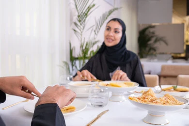 طاولة مليئة باطباق متنوعه ومختلفه من المأكولات الشهية، قضاء وقت ممتعه خارج المنزل في عطلة نهاية الاسبوع, سيدة عربية خليجية اماراتية محجبة ترتدي العباءة تقوم بتناول الطعام في المطعم برفقة زوجها, مفهوم الاسره السعيده