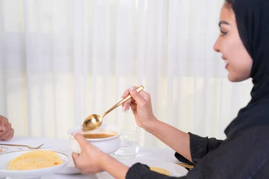 مطاعم راقية في الامارات، طاولة مليئة باطباق متنوعه ومختلفه من المأكولات الشهية، قضاء وقت ممتعه خارج المنزل في عطلة نهاية الاسبوع, صورة من الجانب لامرأة عربية خليجية اماراتية محجبة ترتدي العباءة تقوم بتناول الطعام في المطعم