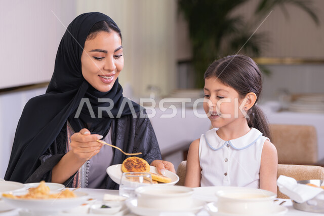 مفهوم الاهتمام والرعاية بالأبناء، أم عربية خليجية اماراتية محجبة ترتدي عباءة سوداء رفقة ابنتها الصغيرة يقومون بتناول الطعام في مطعم فاخر، امرأة عربية خليجية سعودية تحمل بيدها طبق الأكل