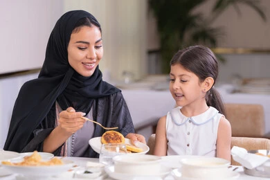 مفهوم الاهتمام والرعاية بالأبناء، أم عربية خليجية اماراتية محجبة ترتدي عباءة سوداء رفقة ابنتها الصغيرة يقومون بتناول الطعام في مطعم فاخر، امرأة عربية خليجية سعودية تحمل بيدها طبق الأكل
