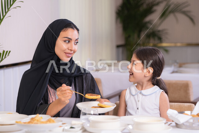قضاء وقت ممتع في مطعم فاخر، مفهوم الاهتمام والرعاية بالأبناء، أم عربية خليجية اماراتية محجبة ترتدي عباءة سوداء رفقة ابنتها الصغيرة يقومون بتناول الطعام في مطعم فاخر، امرأة عربية خليجية سعودية تحمل بيدها طبق الأكل