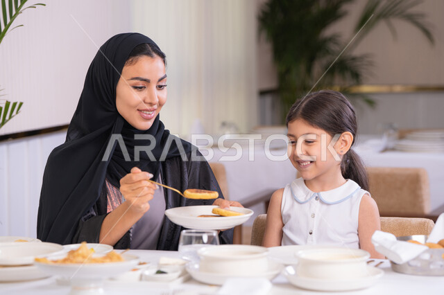 مفهوم الاهتمام والرعاية بالأبناء، أم عربية خليجية اماراتية محجبة ترتدي عباءة سوداء رفقة ابنتها الصغيرة يقومون بتناول الطعام في مطعم فاخر، امرأة عربية خليجية سعودية تحمل بيدها طبق الأكل