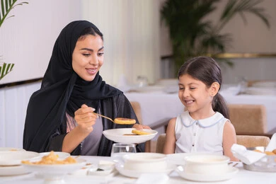 مفهوم الاهتمام والرعاية بالأبناء، أم عربية خليجية اماراتية محجبة ترتدي عباءة سوداء رفقة ابنتها الصغيرة يقومون بتناول الطعام في مطعم فاخر، امرأة عربية خليجية سعودية تحمل بيدها طبق الأكل