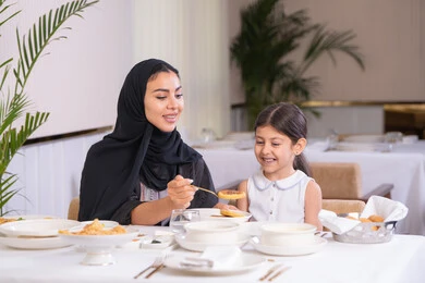 قضاء وقت ممتع في مطعم فاخر، مفهوم الاهتمام والرعاية بالأبناء، أم عربية خليجية اماراتية محجبة ترتدي عباءة سوداء رفقة ابنتها الصغيرة يقومون بتناول الطعام في مطعم فاخر، امرأة عربية خليجية سعودية تحمل بيدها طبق الأكل