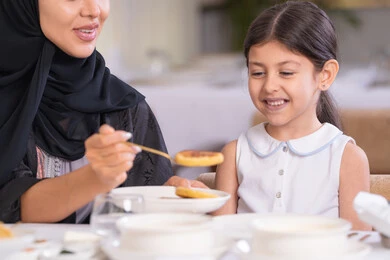 تعابير السعادة والسرور، قضاء وقت ممتع خارج المنزل، مفهوم الاهتمام والرعاية بالأبناء، صورة مقربة لأم عربية خليجية اماراتية محجبة ترتدي عباءة سوداء رفقة ابنتها الصغيرة يقومون بتناول الطعام في مطعم فاخر، امرأة عربية خليجية سعودية تحمل بيدها طبق الأكل