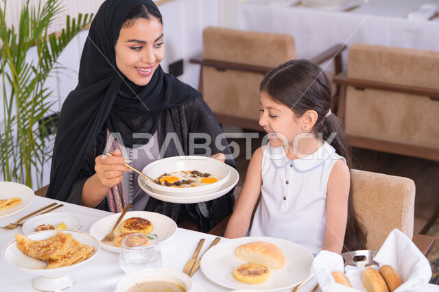 ايماءات السعادة والسرور، قضاء وقت ممتع خارج المنزل، مفهوم الاهتمام والرعاية بالأبناء، أم عربية خليجية اماراتية محجبة ترتدي عباءة سوداء رفقة ابنتها الصغيرة يقومون بتناول الطعام في مطعم فاخر، امرأة عربية خليجية سعودية تحمل بيدها طبق الأكل