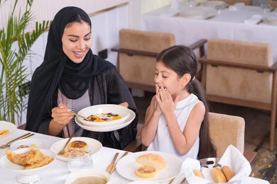 ايماءات السعادة والسرور، قضاء وقت ممتع خارج المنزل، مفهوم الاهتمام والرعاية بالأبناء، أم عربية خليجية اماراتية محجبة ترتدي عباءة سوداء رفقة ابنتها الصغيرة يقومون بتناول الطعام في مطعم فاخر، امرأة عربية خليجية سعودية تحمل بيدها طبق الأكل