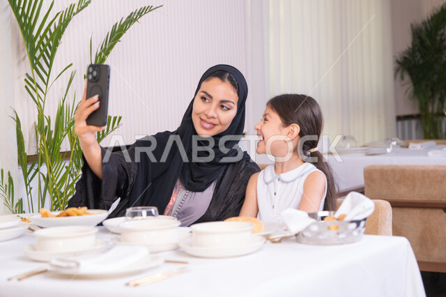 Documenting happy moments, spending quality time outdoors in a high-end restaurant, an Emirati Gulf Arab mother and her daughter taking a selfie, a veiled Saudi Gulf Arab woman wearing a black abaya holding a mobile phone, enjoying the weekend