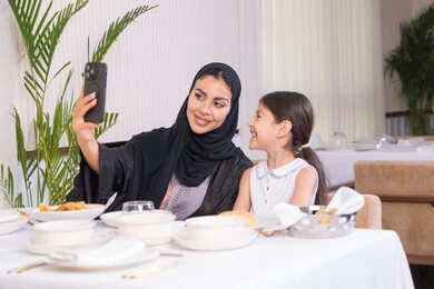 توثيق اللحظات السعيدة، قضاء وقت ممتع خارجي في احدى المطاعم الراقية، أم عربية خليجية اماراتية رفقة ابنتها تقوم بالتقاط صورة سيلفي، امرأة عربية خليجية سعودية محجبة ترتدي عباءة سوداء تحمل بيددها الهاتف الجوال، الاستمتاع في عطلة نهاية الأسبوع