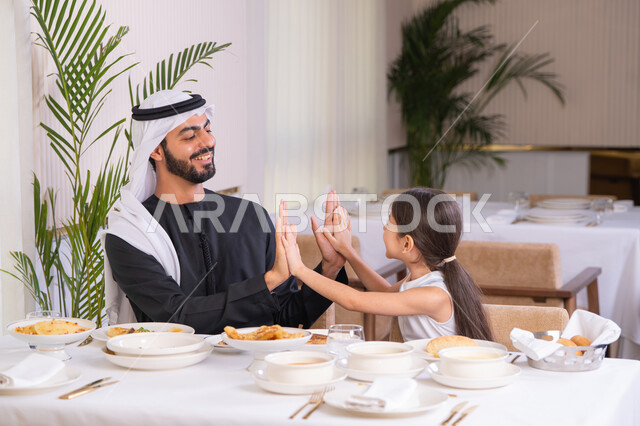 دفء العائلة وقيمة الترابط الأسري، جلسة عائلية في احدى المطاعم الراقية، اب عربي خليجي اماراتي يرتدي الكندورة والغترة يلاعب طفلته بإيماءات المحبة والفرح، التسلية والترفيه في عطلة نهاية الاسبوع
