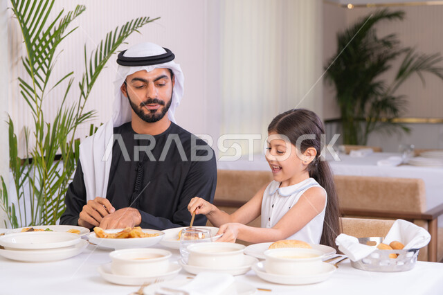 Gestures of happiness and joy, supporting the child's independence with a spirit of fun, an Arab Gulf Emirati father wearing a kandura and ghutra enjoying spending time with his daughter in a high-end restaurant, the concept of care and attention, a table full of delicious Western food dishes, entertainment and recreation on holidays