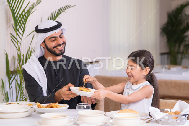 ايماءات السعادة و السرور، دعم استقلالية الطفلة بتعابير المرح، اب عربي خليجي اماراتي يرتدي الكندورة والغترة يستمتع بقضاء وقت مع ابنته في مطعم راقي، مفهوم الرعاية والاهتمام، طاولة مليئة بأطباق الأطعمة الغربية الشهية، التسلية والترفيه في العطلة