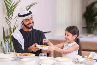 ايماءات السعادة و السرور، دعم استقلالية الطفلة بتعابير المرح، اب عربي خليجي اماراتي يرتدي الكندورة والغترة يستمتع بقضاء وقت مع ابنته في مطعم راقي، مفهوم الرعاية والاهتمام، طاولة مليئة بأطباق الأطعمة الغربية الشهية، التسلية والترفيه في العطلة