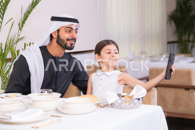 الاهتمام ودعم الابناء، التسلية والترفيه في العطلات، جلسة عائلية دافئة، فتاه عربية خليجية اماراتية ترتدي الزي الكاجوال تجلس مع والدها في المطعم تقوم بالتقاط صورة سيلفي تذكارية للحظات السعيدة بإيماءات السعادة والسرور