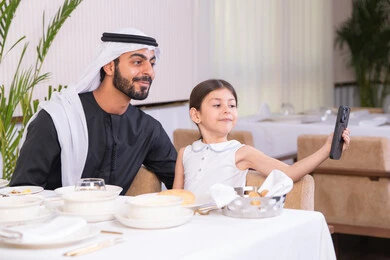 الاهتمام ودعم الابناء، التسلية والترفيه في العطلات، جلسة عائلية دافئة، فتاه عربية خليجية اماراتية ترتدي الزي الكاجوال تجلس مع والدها في المطعم تقوم بالتقاط صورة سيلفي تذكارية للحظات السعيدة بإيماءات السعادة والسرور