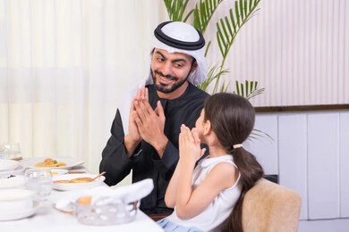 مفهوم الرعاية والاهتمام بالأبناء، الاستمتاع مع افراد الأسره، اب عربي خليجي اماراتي يرتدي الكندورة والغترة يقوم بتناول الطعام مع ابنته في مطعم راقي، طاولة مليئة بأطباق الأطعمة الغربية، التسلية والترفيه في العطلة، ايماءات السعادة و السرور