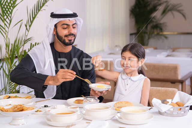 مفهوم الرعاية والاهتمام بالأبناء، الاستمتاع مع افراد الأسره، اب عربي خليجي اماراتي يرتدي الكندورة والغترة يقوم بتناول الطعام مع ابنته في مطعم راقي، طاولة مليئة بأطباق الأطعمة الغربية، التسلية والترفيه في العطلة، ايماءات السعادة و السرور