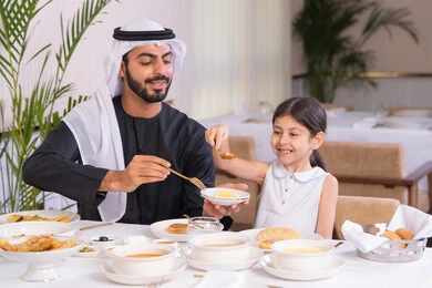 مفهوم الرعاية والاهتمام بالأبناء، الاستمتاع مع افراد الأسره، اب عربي خليجي اماراتي يرتدي الكندورة والغترة يقوم بتناول الطعام مع ابنته في مطعم راقي، طاولة مليئة بأطباق الأطعمة الغربية، التسلية والترفيه في العطلة، ايماءات السعادة و السرور