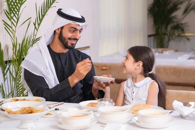 مفهوم الرعاية والاهتمام بالأبناء، الاستمتاع مع افراد الأسره، اب عربي خليجي اماراتي يرتدي الكندورة والغترة يقوم بتناول الطعام مع ابنته في مطعم راقي، طاولة مليئة بأطباق الأطعمة الغربية، التسلية والترفيه في العطلة، ايماءات السعادة و السرور