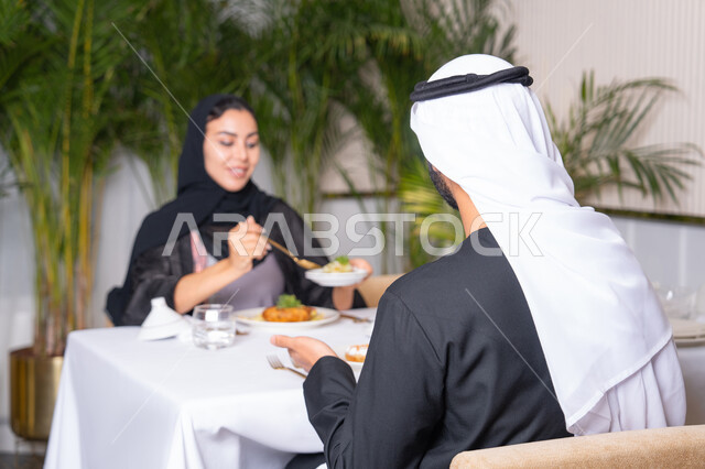 المحبة والمودة بين الزوجين، مطعم راقي في الامارات العربية المتحدة، طاولة مليئة باطباق متنوعه ومختلفه من المأكولات الشهية، قضاء وقت ممتعه خارج المنزل في عطلة نهاية الاسبوع, زوجان عربيان اماراتيان يقوم بتناول الطعام في المطعم, مفهوم الاسره السعيده