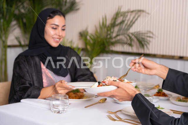Gestures of happiness and pleasure, a table full of various delicious dishes, spending a pleasant time outside the home on the weekend, a veiled Emirati Gulf Arab woman wearing an abaya eating at a restaurant, the concept of a happy family
