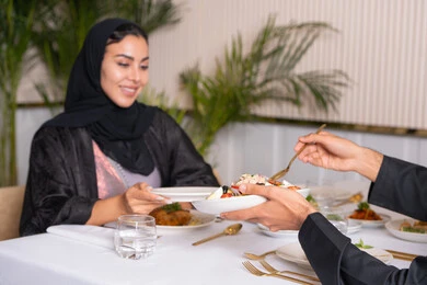 Gestures of happiness and pleasure, a table full of various delicious dishes, spending a pleasant time outside the home on the weekend, a veiled Emirati Gulf Arab woman wearing an abaya eating at a restaurant, the concept of a happy family