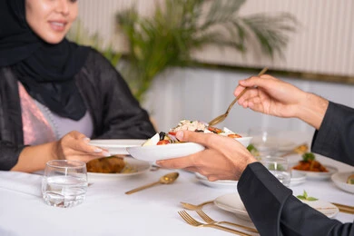 Expressions of happiness and pleasure, a table full of various delicious dishes, spending a pleasant time outside the home on the weekend, a close-up photo of a veiled Emirati Gulf Arab woman wearing an abaya eating in a restaurant with her husband, the concept of a happy family